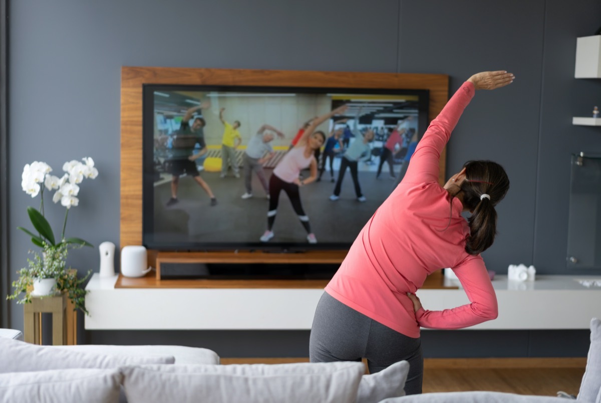 Back view of senior woman following an online stretching class looking at TV screen.