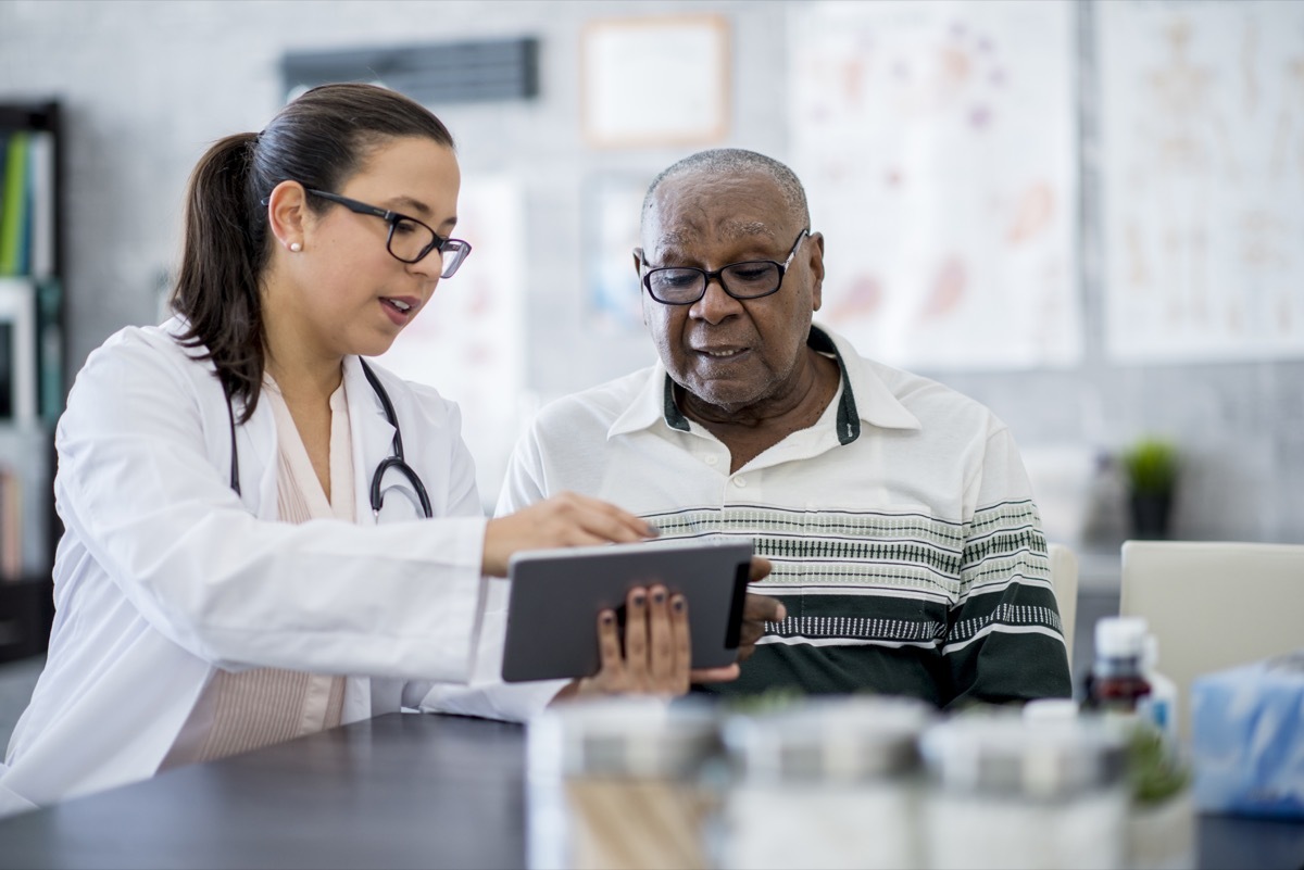 A senior man is indoors in a hospital room. He is watching his female doctor using a tablet computer. She is explaining a medication schedule to him.