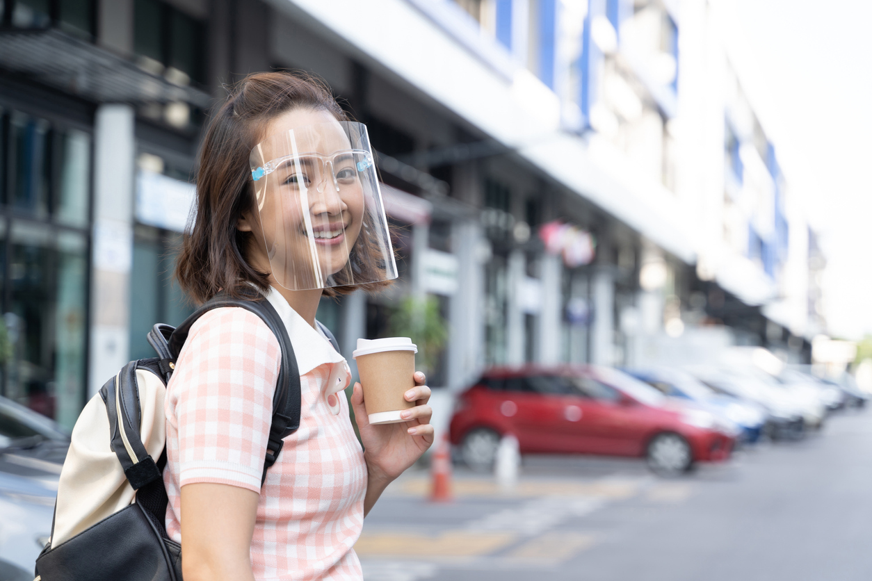 A young Asian woman smiles while wearing a face mask and holding a cup of coffee