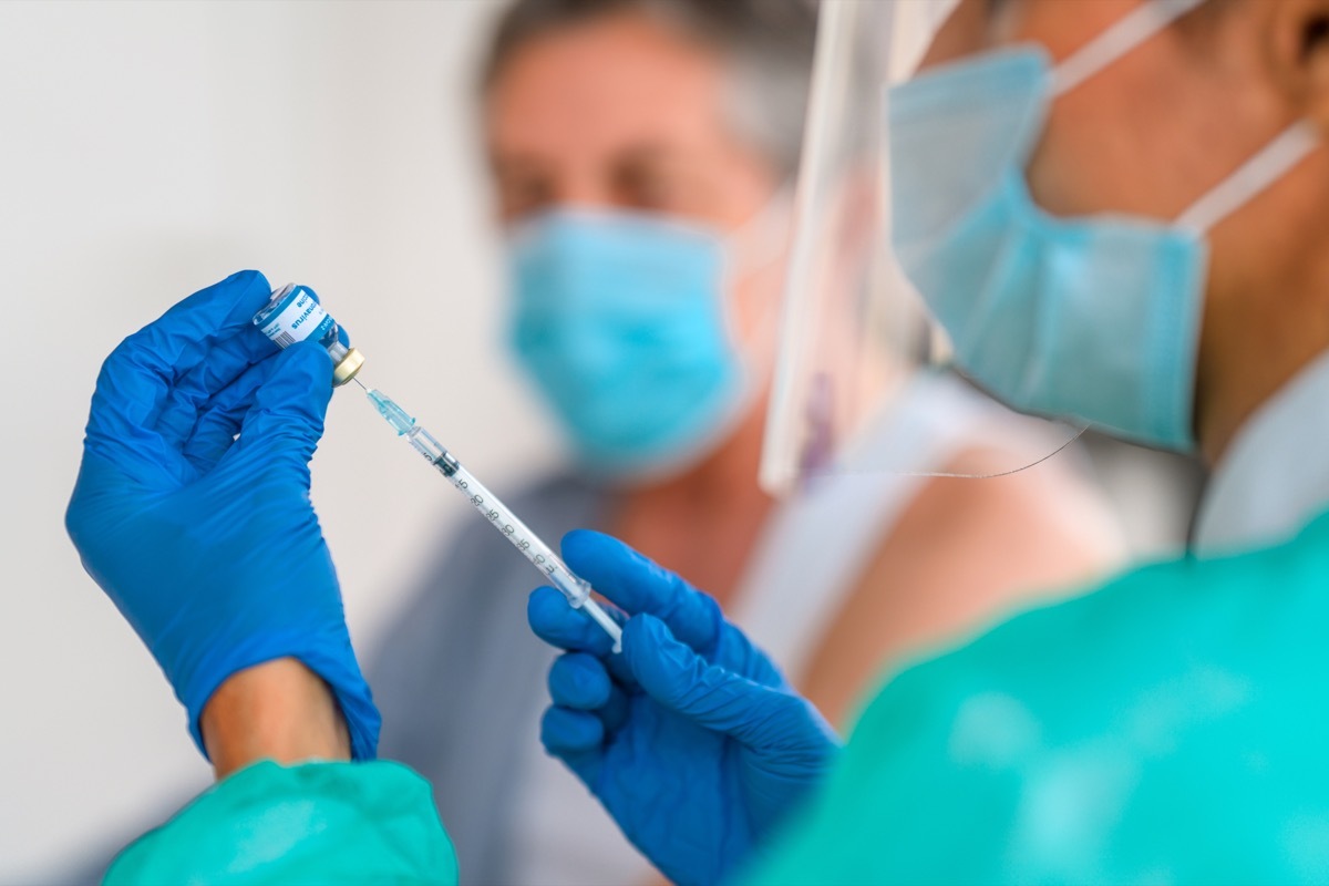 doctor is sitting in a doctor's office and is filling syringe for Covid-19 vaccine. She is wearing protective gloves, protective coat, face shield and protective face mask.