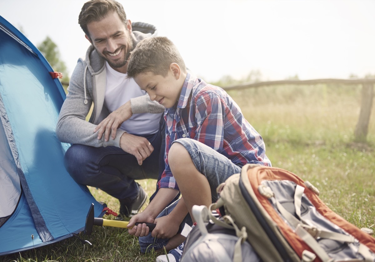 father watching son hammer nail to put up a tent outside