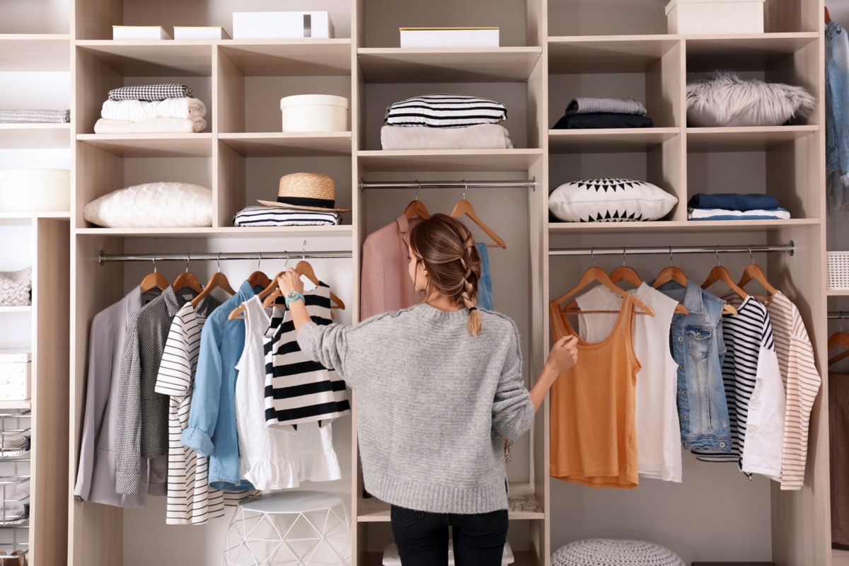 Woman choosing outfit from large wardrobe closet with stylish clothes and home stuff