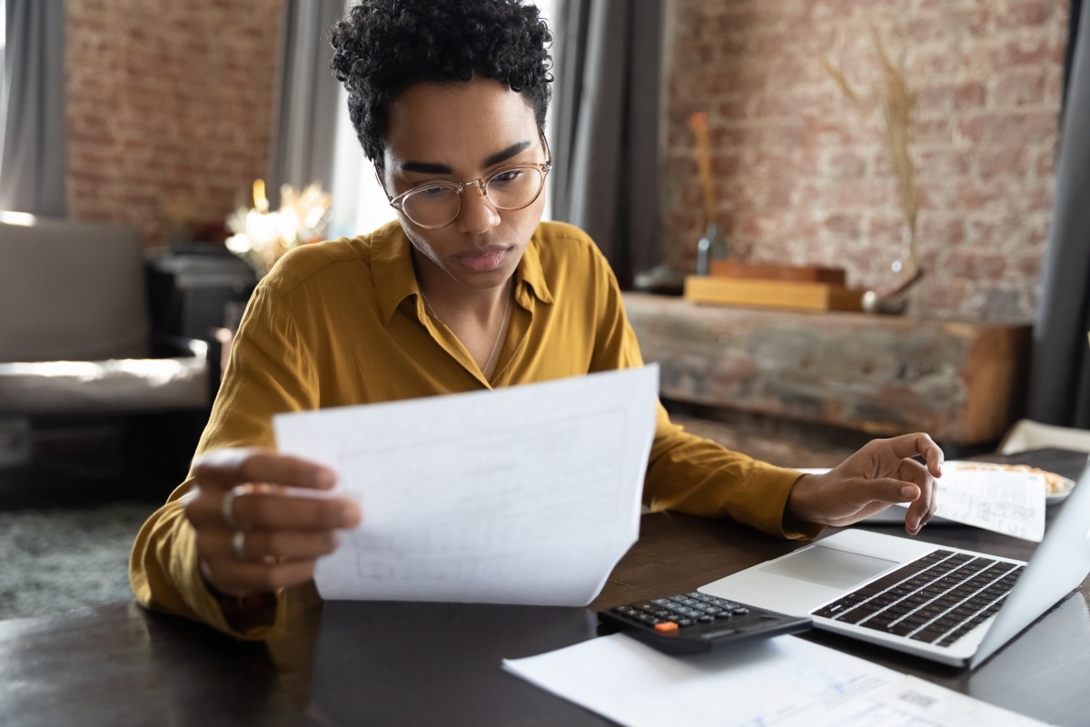 Focused young woman in eyeglasses looking through paper documents with laptop and calculator