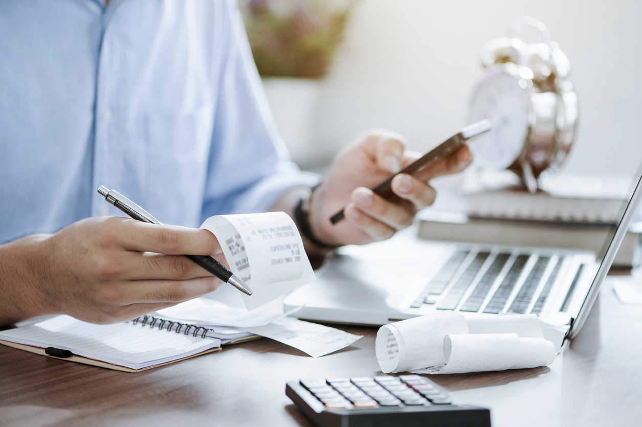 Close up of a person holding receipts and their phone while filing their taxes using a laptop and calculator