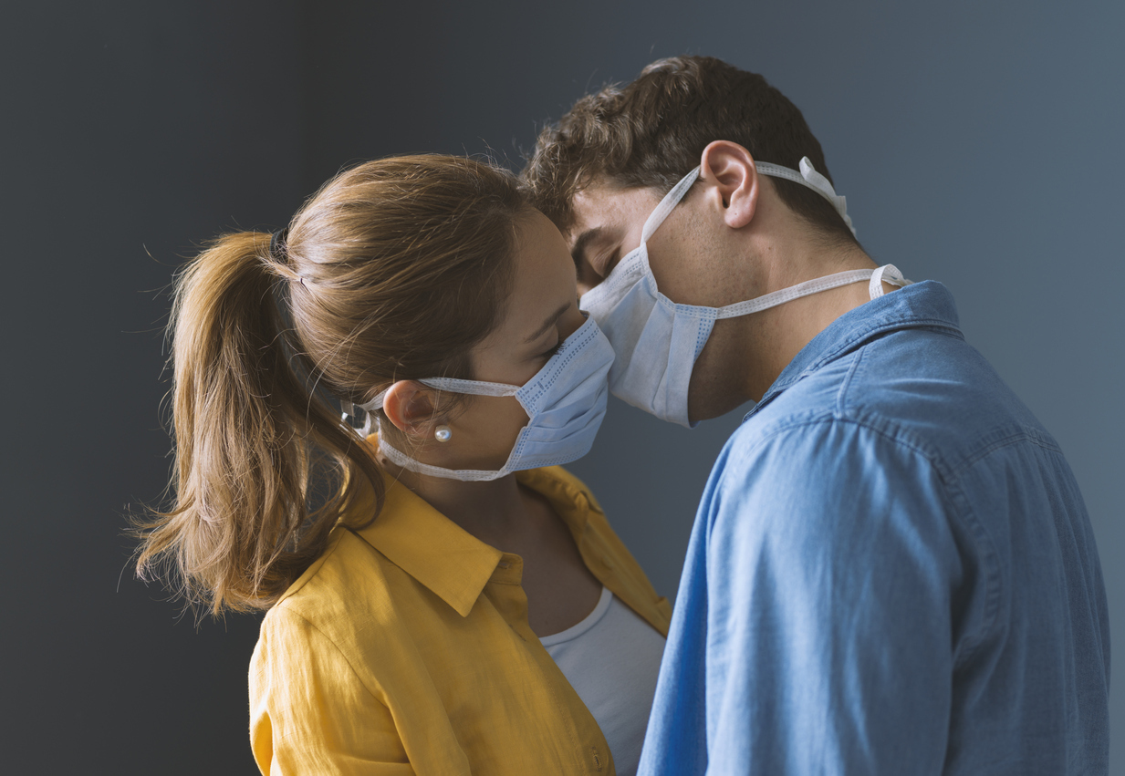 A young male and female couple lean in to kiss each other while both wearing face masks.