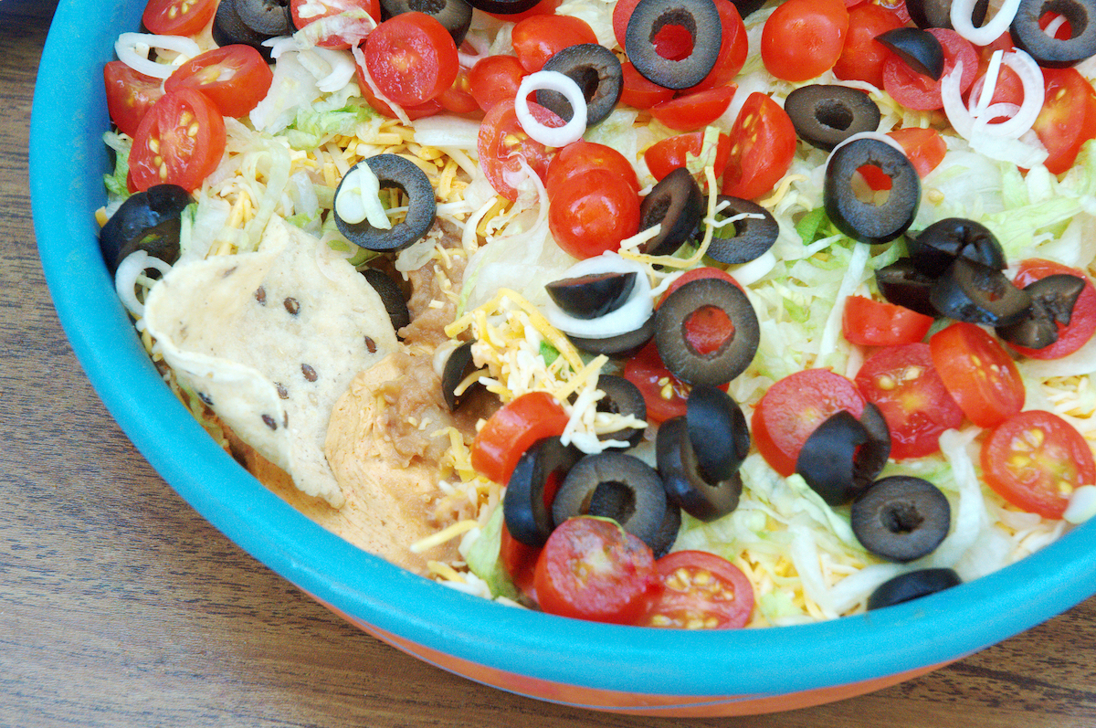 layered taco dip in blue bowl with olives, tomatoes, cheese, lettuce