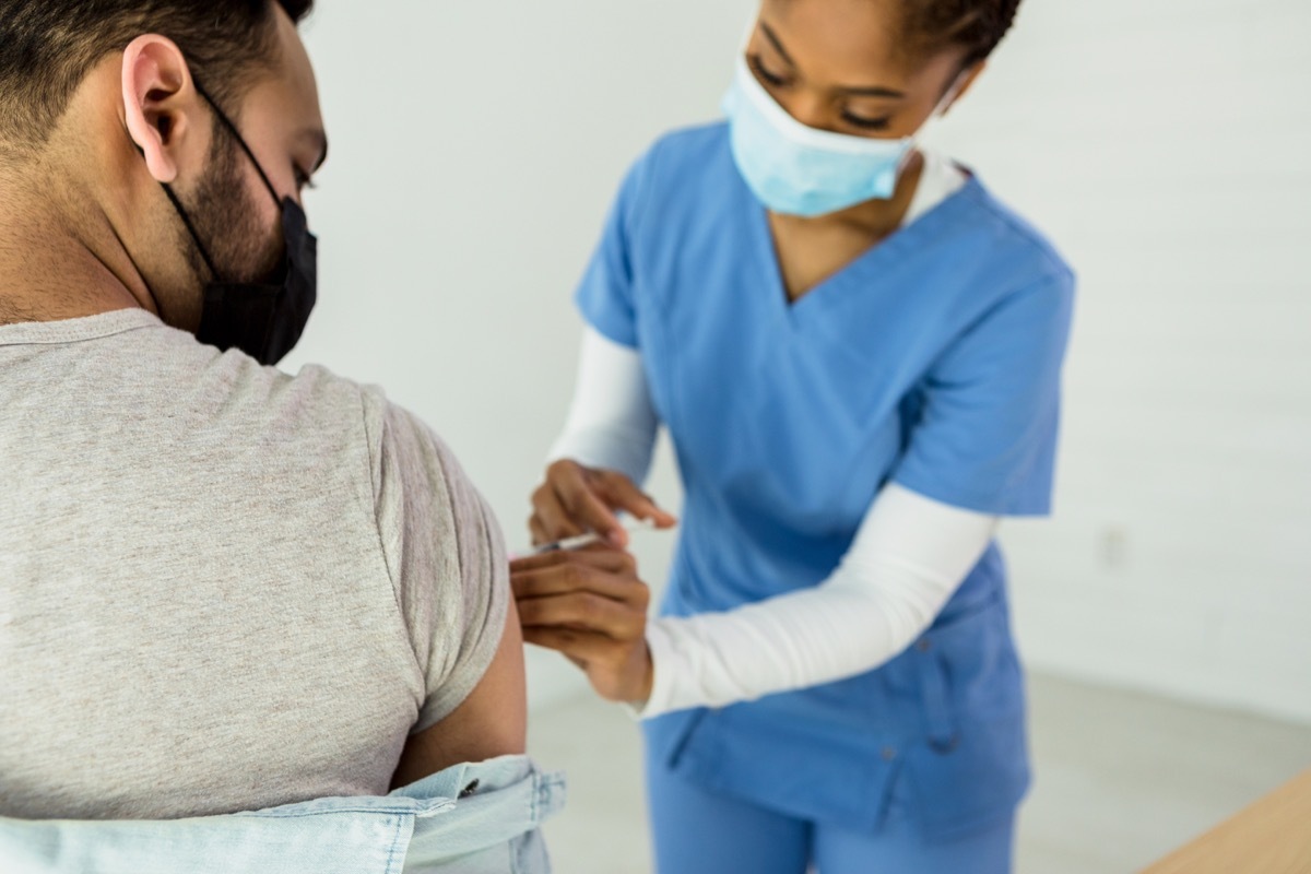 The mid adult female nurse administers the COVID-19 vaccine into the unrecognizable mid adult male patient's upper arm.