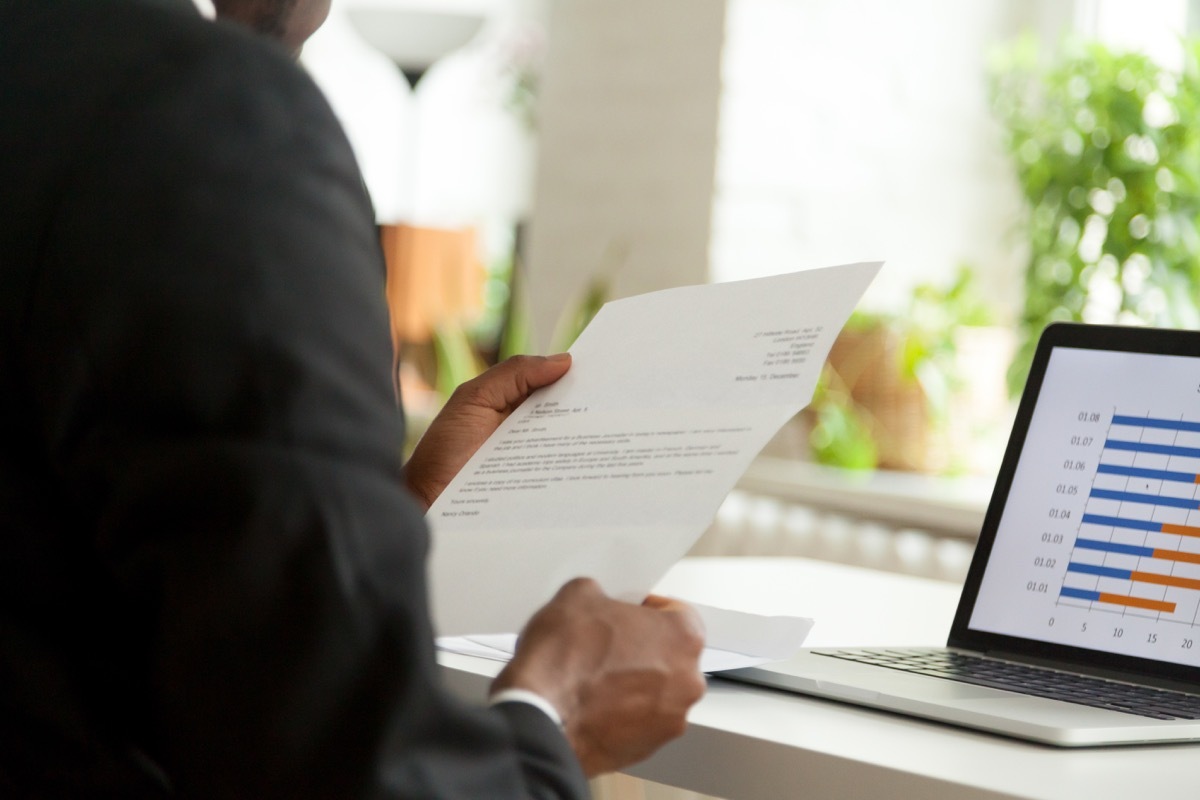 businessman in a black suit holding a letter while seated in front of his laptop showing a graph