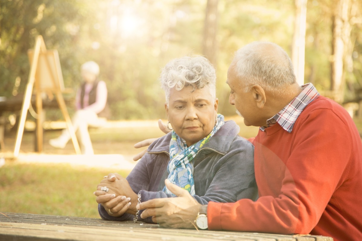 Senior adult couple talk at outdoor park in spring or autumn season. They sit on a park bench and discuss their relationship difficulties.