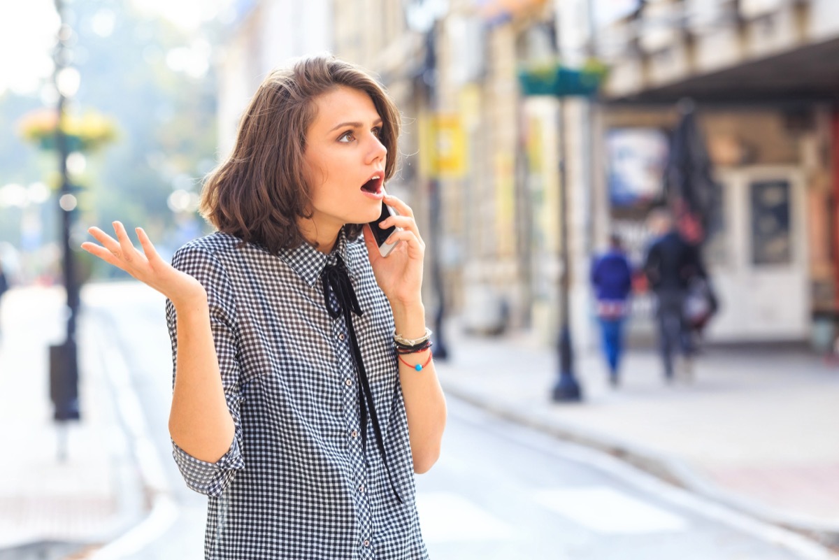 Angry woman talking on smart phone on street. Wears casual clothes.