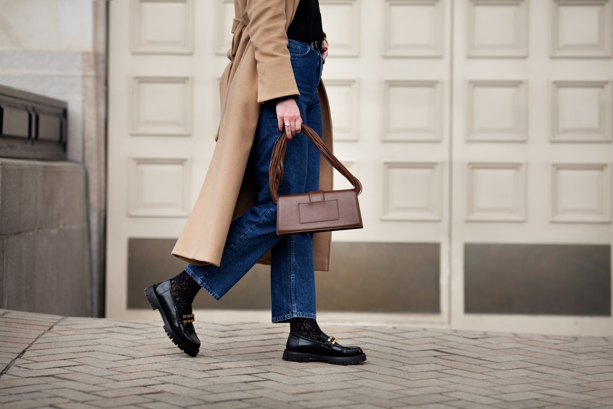 Side view woman walking street in fashionable spring or autumn clothes cashmere coat, jeans, black loafers shoes and handbag. Female model in motion, street style fashion, close up legs