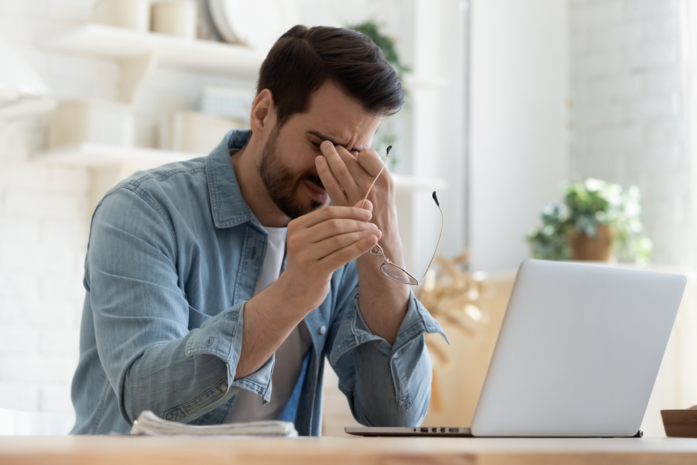 A young man working at a laptop takes off his glasses to rub his eyes with a sleepy, fatigued look