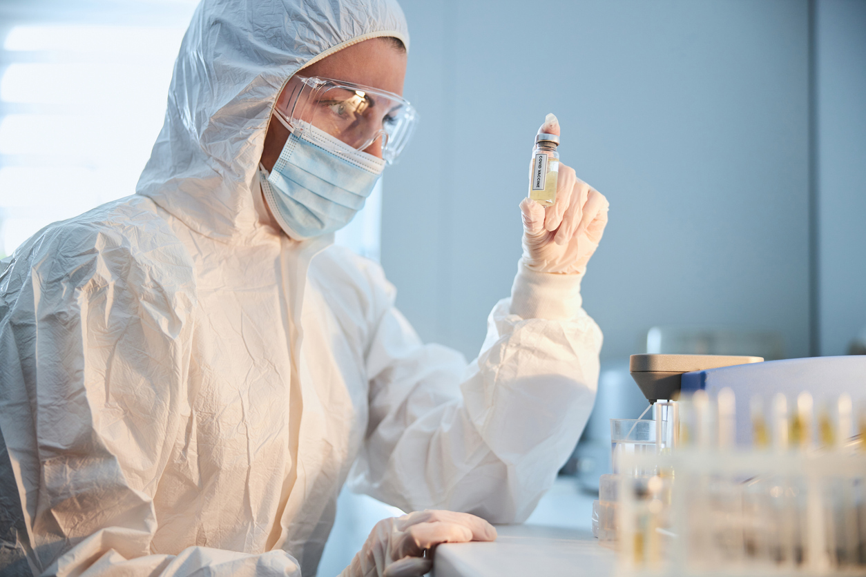 A scientist in a lab wearing full protective gear while looking at a vial of COVID-19 vaccine