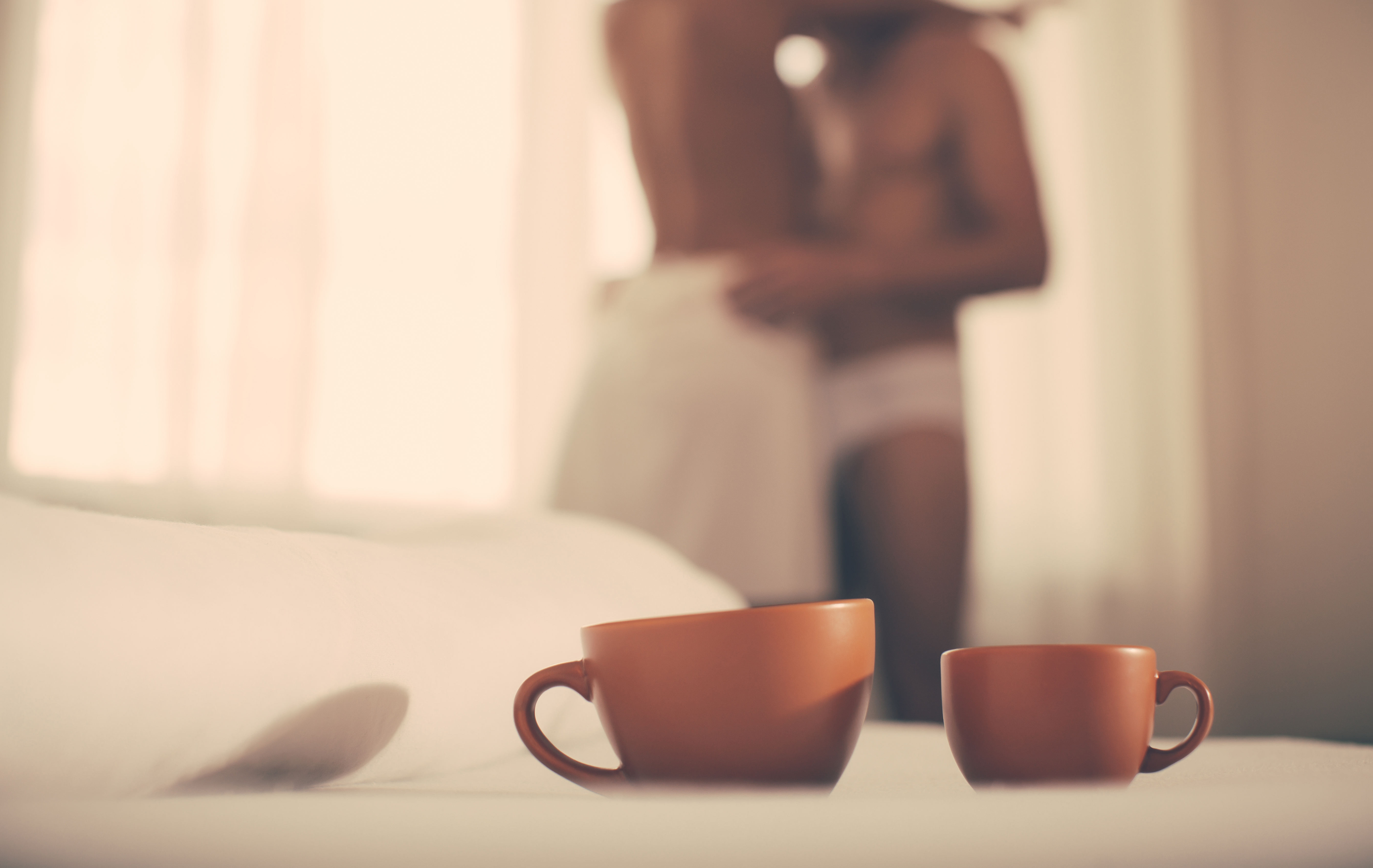 Half-dressed man and woman embracing in the background with two coffee cups in the foreground.