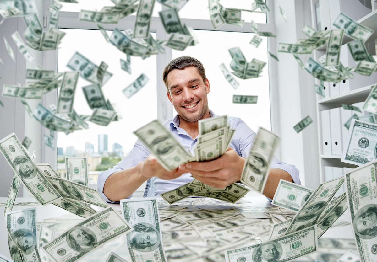 A rich young man sitting at his desk counting money with it flying all around him.