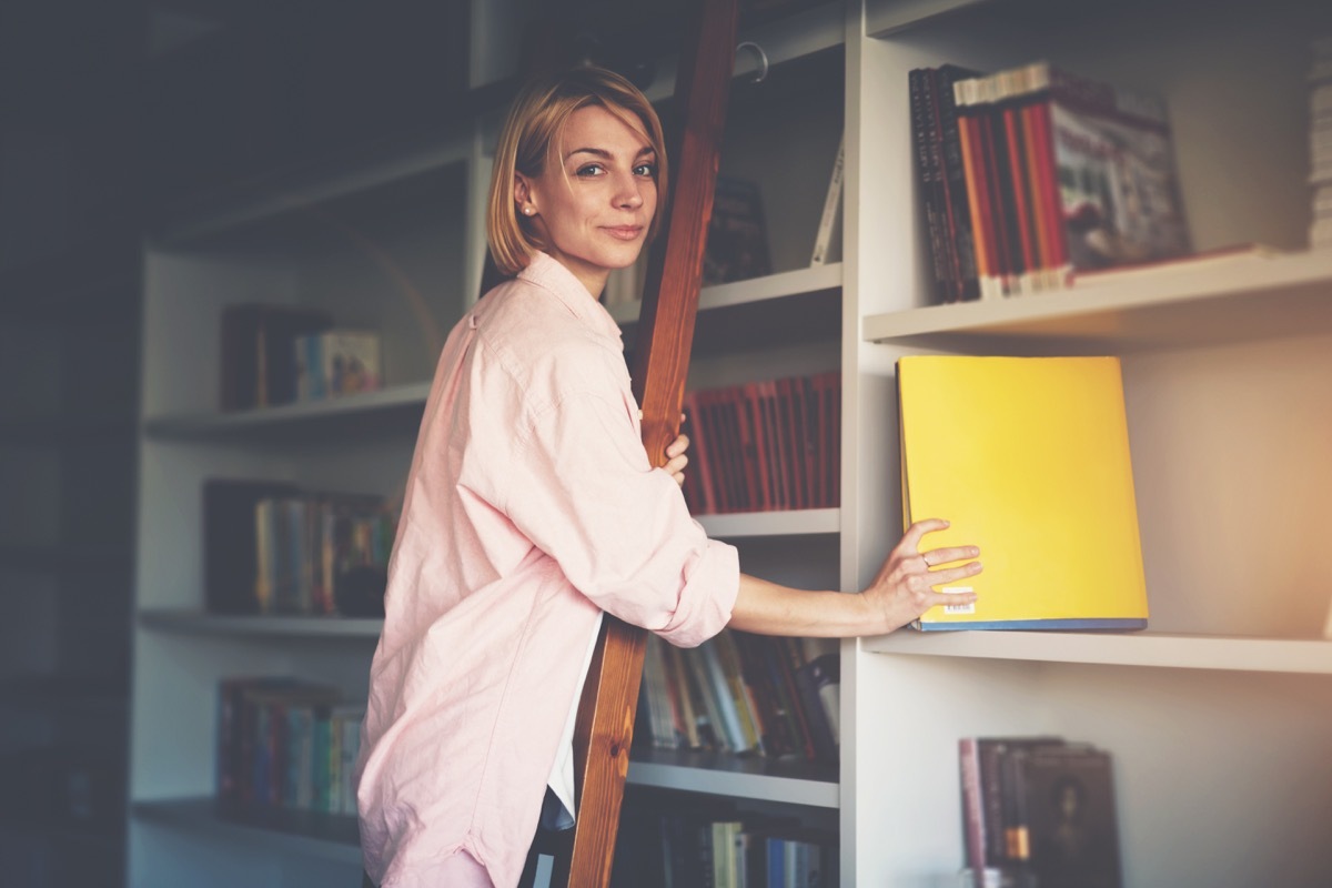 woman picking book from bookshelf toss these things from your house for instant happiness