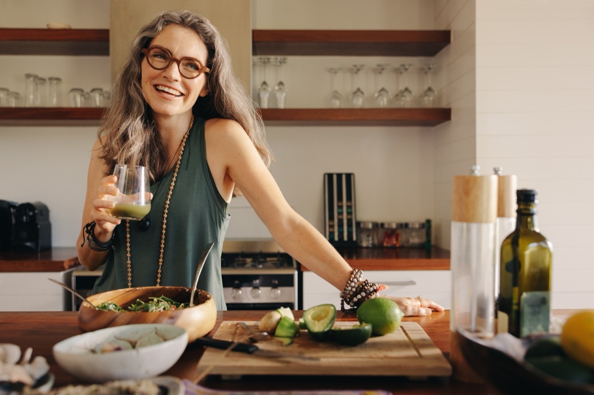 Healthy senior woman smiling while holding some green juice in her kitchen. Mature woman serving herself wholesome vegan food at home. Woman taking care of her aging body with a plant-based diet.