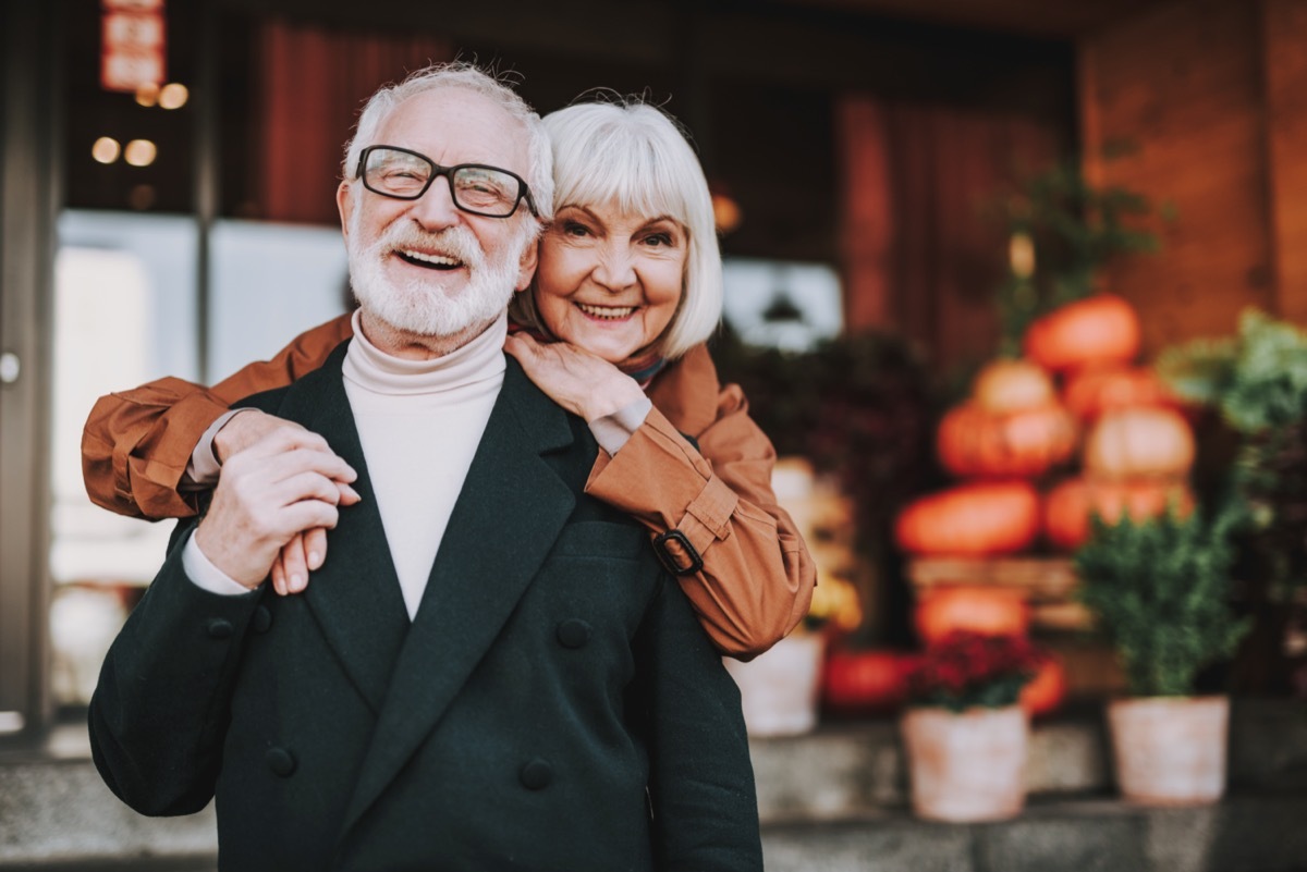 Waist up portrait of happy lady hugging husband from behind while he holding her hand. They looking at camera and smiling