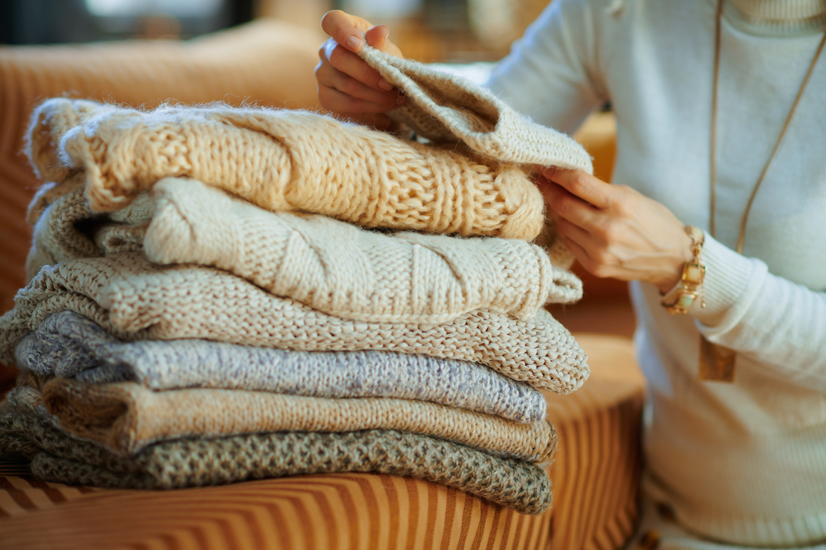 Closeup on trendy woman in white sweater and skirt at modern home in sunny winter day sitting near couch checking sweaters for moth larvae or other damage.