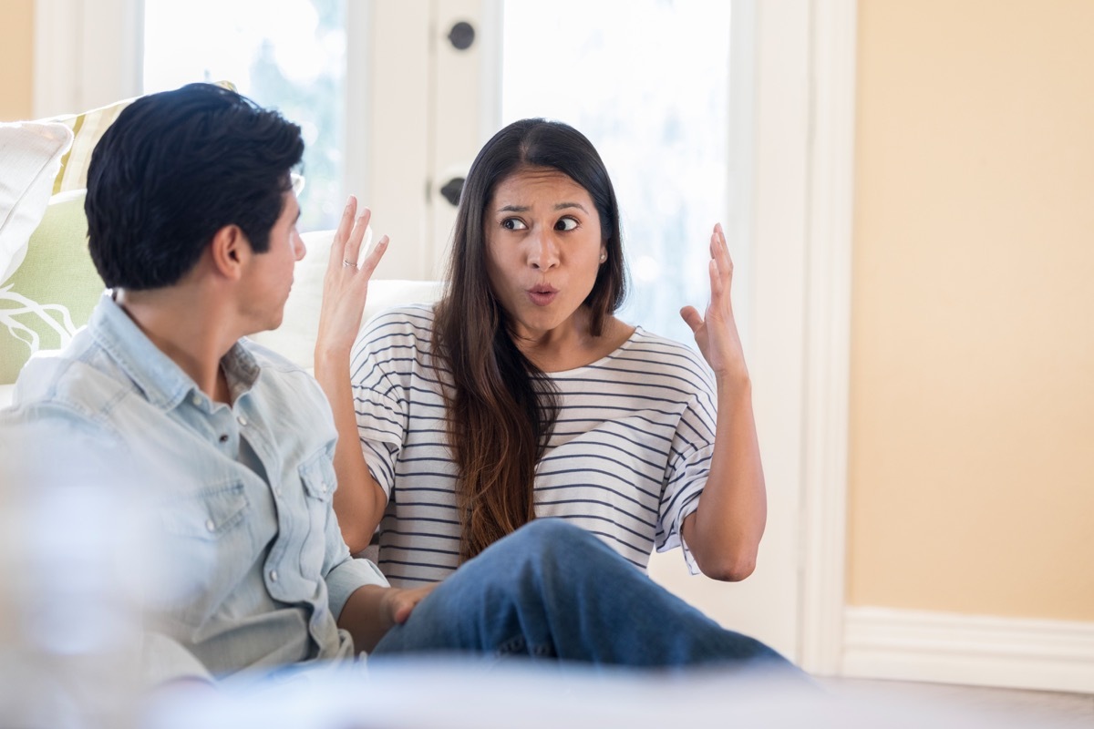 An animated mid adult woman throws up her hands in frustration as she speaks to her unrecognizable husband at home. They are sitting on their living room floor.