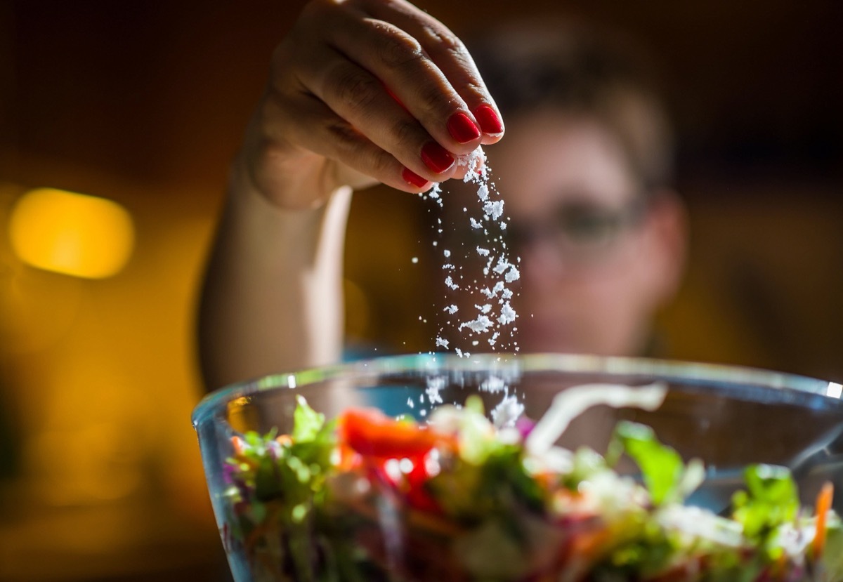 Woman preparing healthy salad in kitchen, adding salt to the bowl