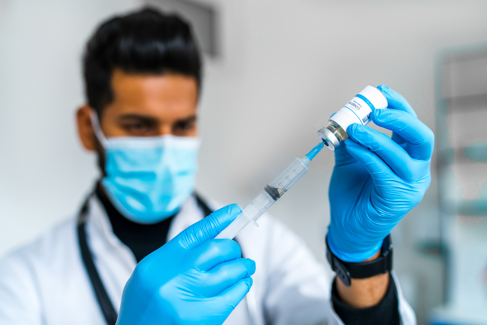 A young medical worker wearing a face mask and gloves fills a vial with COVID-19 vaccine