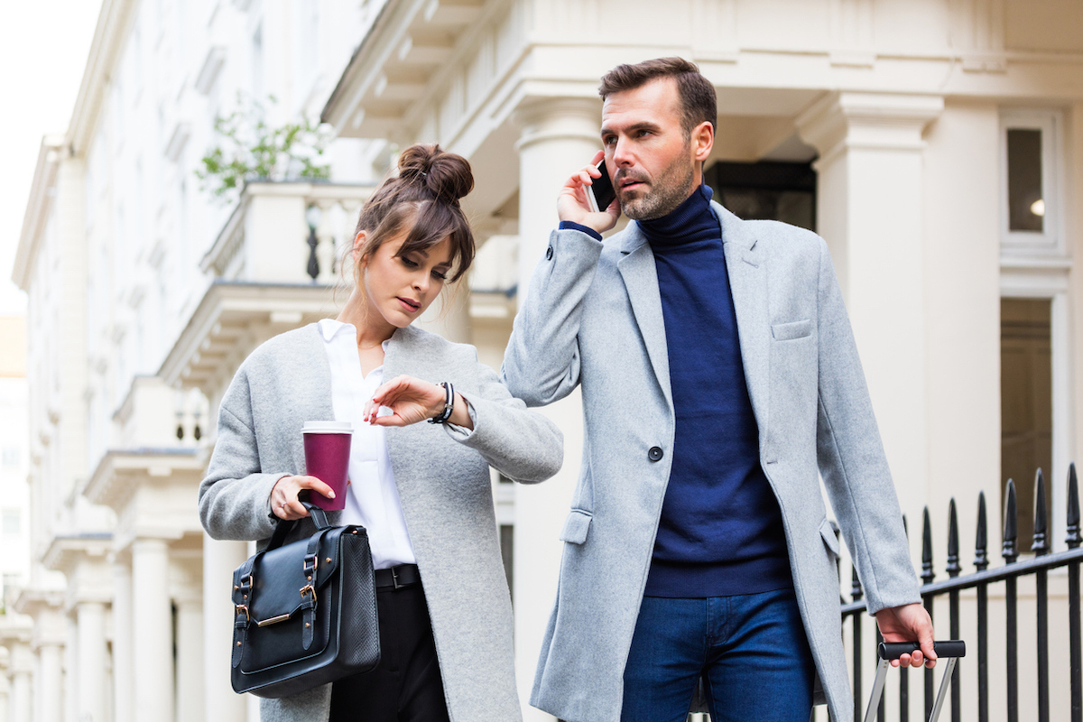 Busy couple walking in the city street, woman holding take away coffee and looking at her watch while man is talking on mobile phone