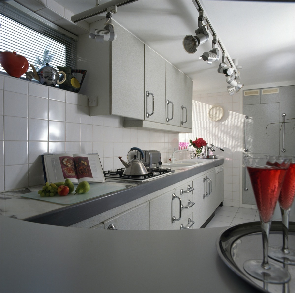 Track spotlights and white tiling in a nineties kitchen with pale gray fitted units and an Alessi kettle on the hob