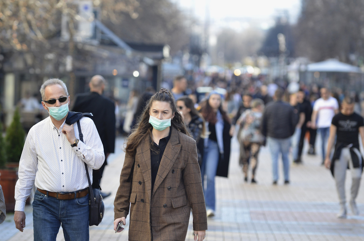 A young woman and an older man are walking on street wearing face masks while others are not