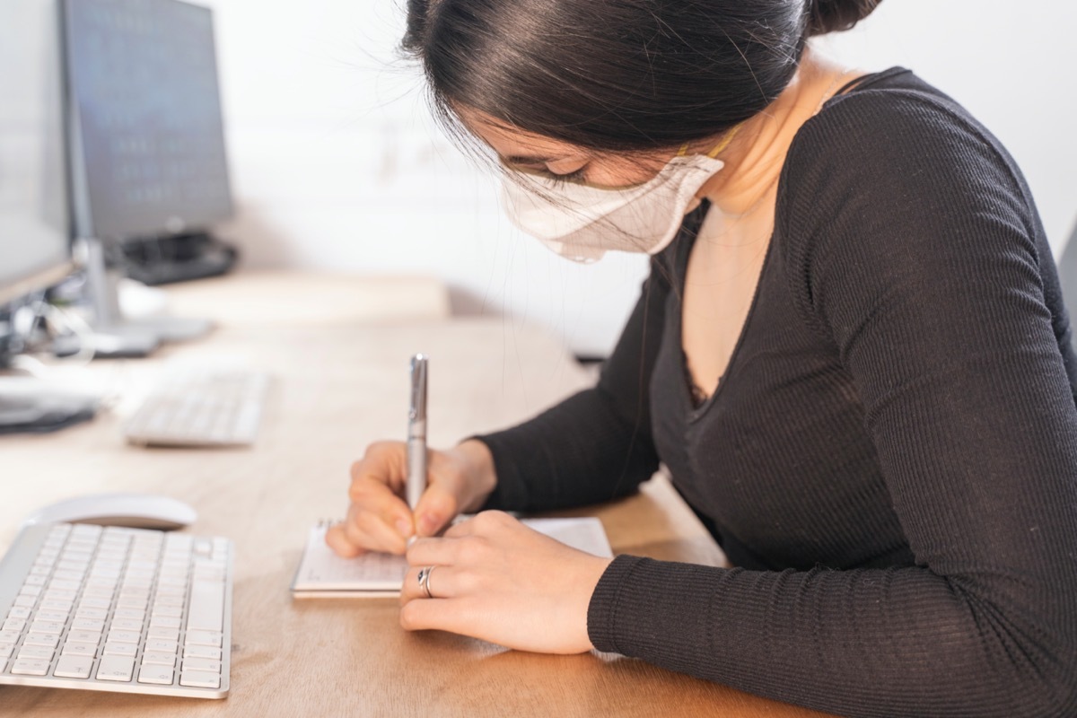 Woman sitting at desk and writing in notebook while wearing mask