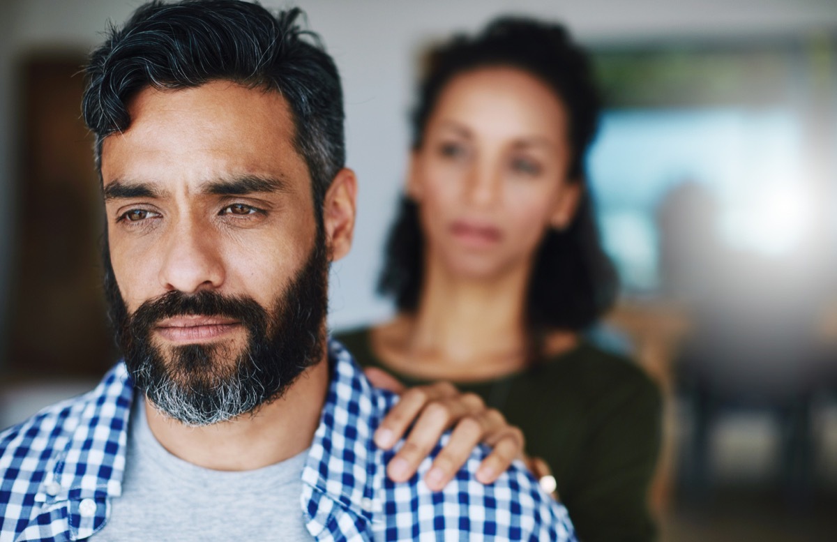 man looking sad while woman puts her hand on his shoulder from behind