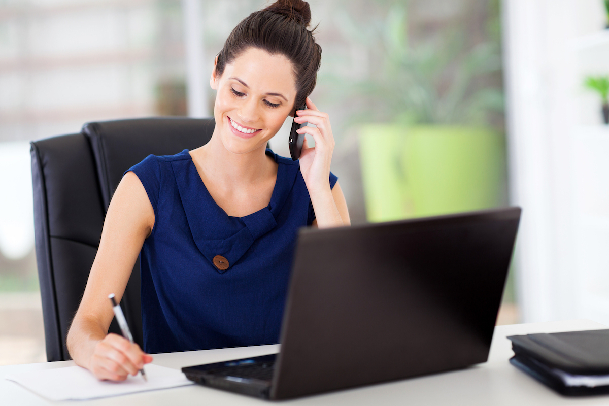 A young woman at her office desk talking on the phone and taking notes while wearing a navy blue dress