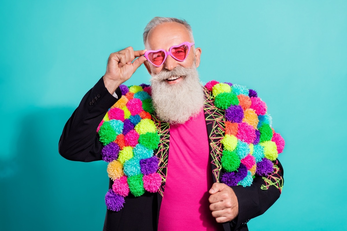 Older man in a colorful jacket and pink heart-shaped sunglasses against a teal background