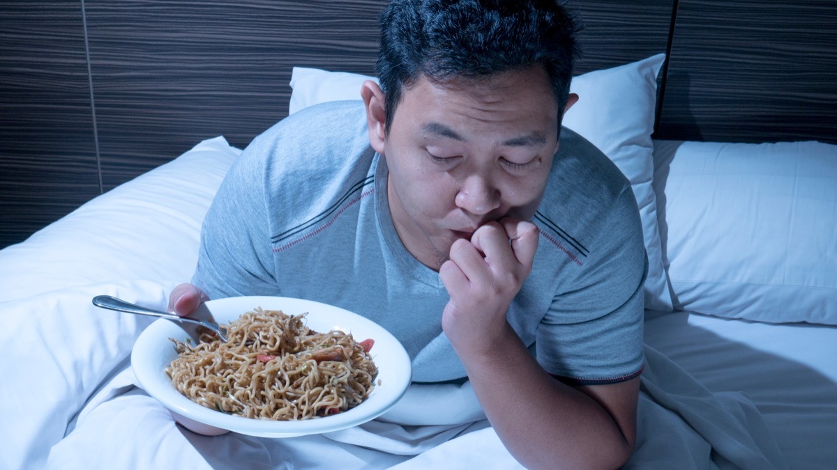 Man sits up in bed late at night with plate of noodles