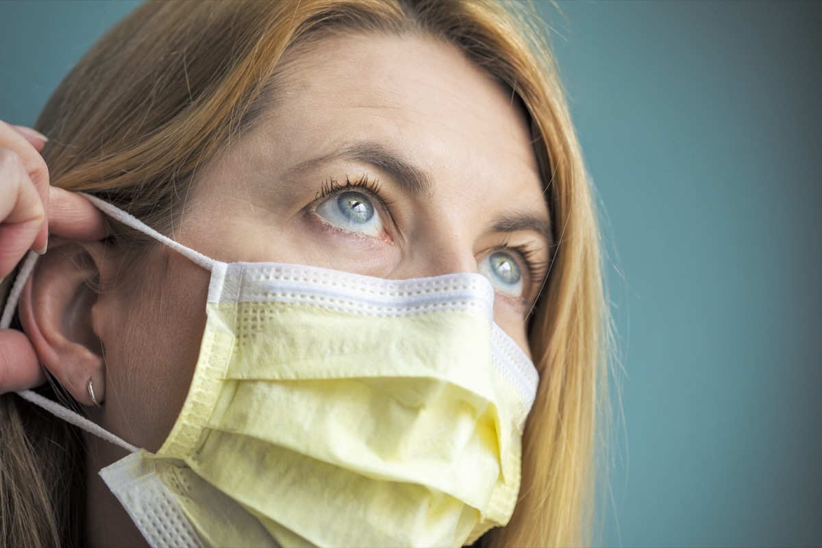 A close up of a woman in her 40's fearful of catching a virus and wearing a face protection mask with her fingers touching it.