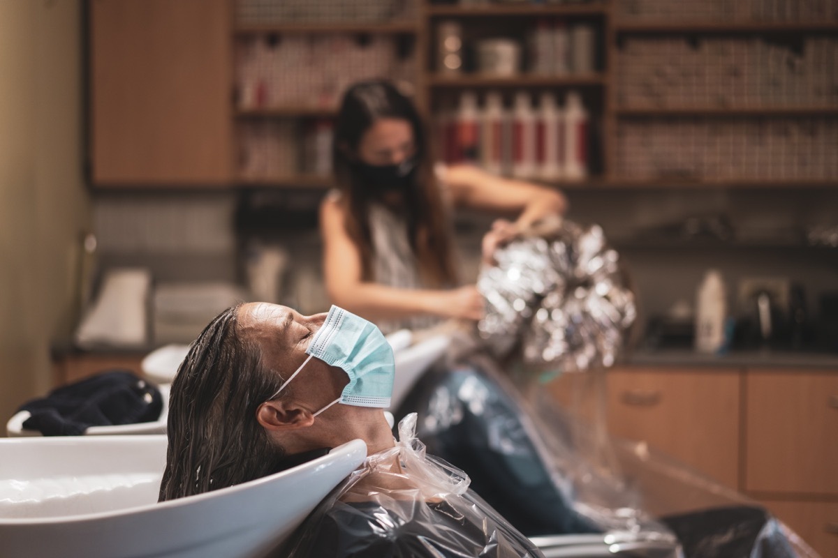 mature adult woman with mouth nose mask and washed hairs relaxing with eyes closed in hairdresser salon during first weeks of reopening after long periode of coronavirus lockdown, coiffeur working on other client with enough social distance blurred in background