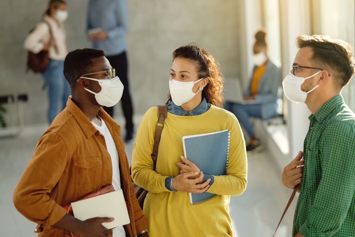 three friends in brightly colored shirts standing in school building wearing masks and holding notebooks