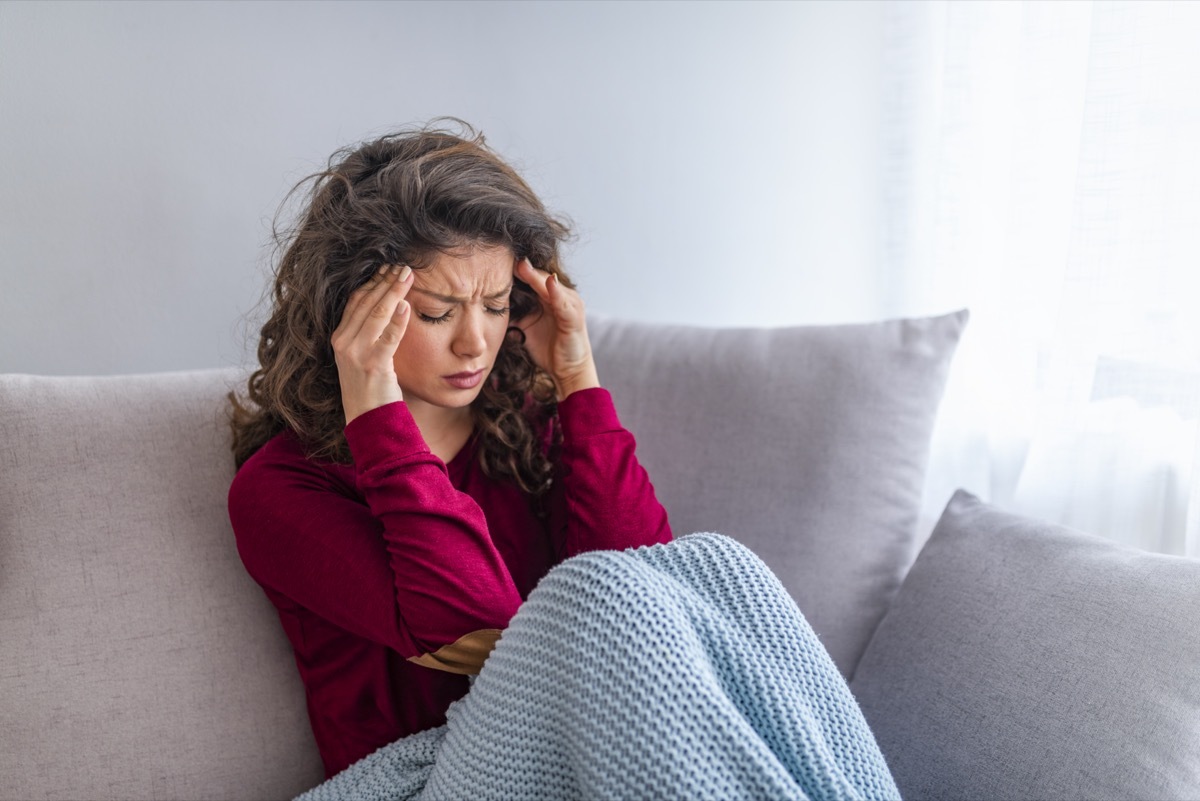 Young woman sitting on a couch, holding her head, having a strong headache. Close up Portrait of young woman with headache.