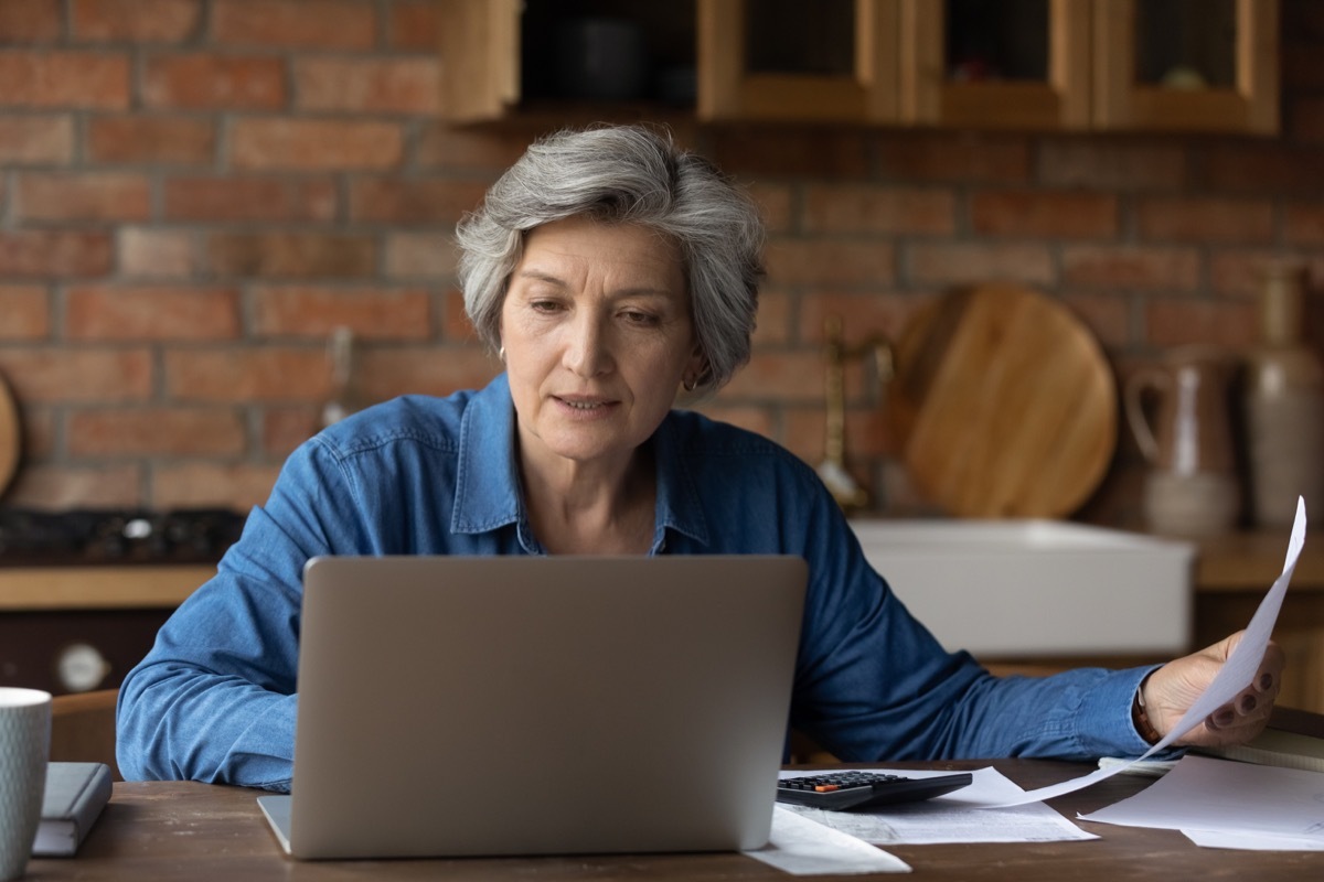 Smiling mature woman using laptop, online baking service, checking financial documents at home, senior grey haired female sitting at table with domestic bills and calculator, accounting