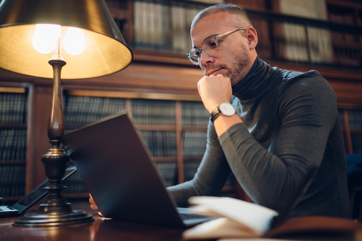 An intellectual man wearing glasses and a gray turtleneck on his laptop at the library