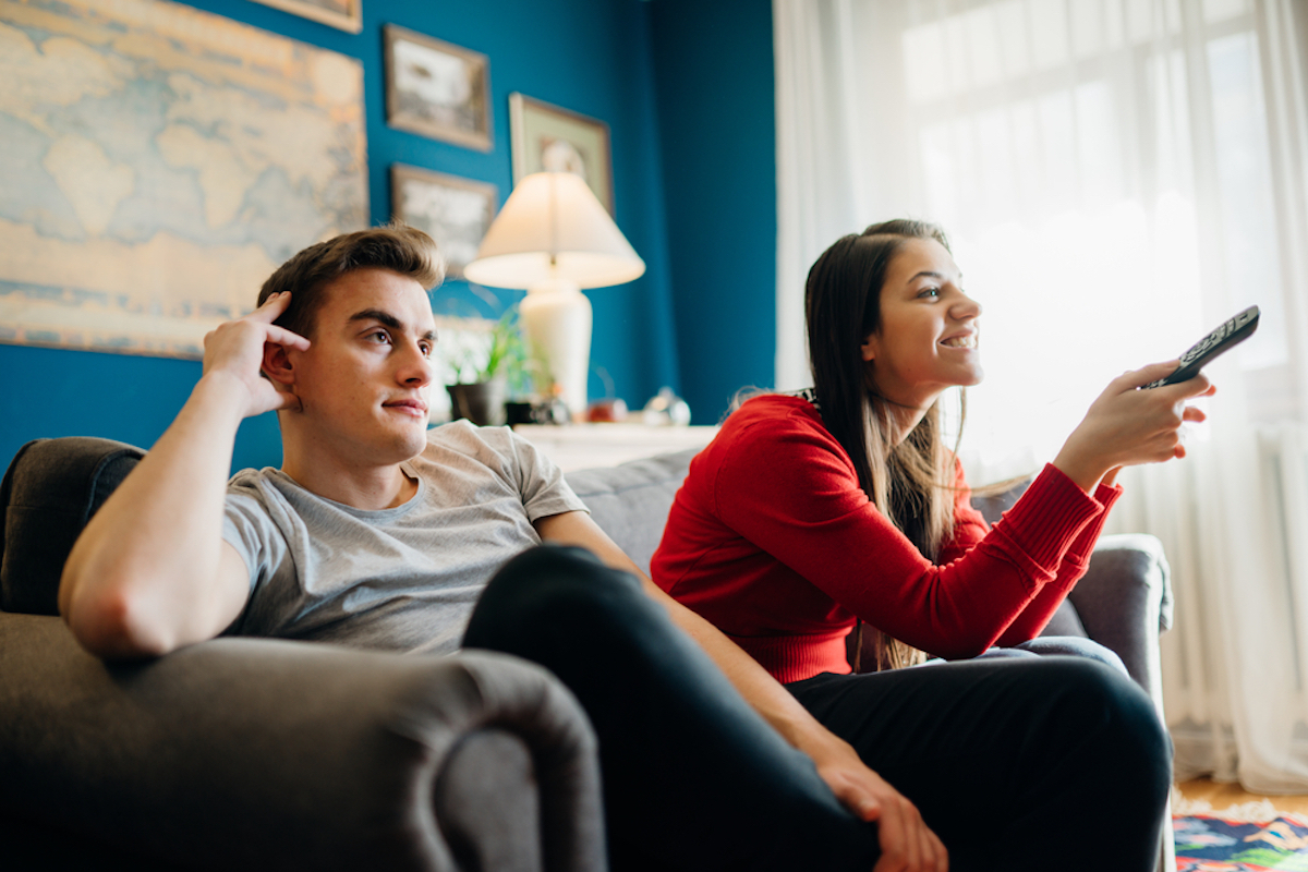 young couple watching tv, young man looks bored while young woman holds remote, smiling